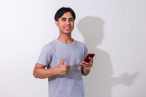 istock Smiling young Asian man showing thumbs up while holding a smartphone, isolated on white background. Perfect for promotion, mobile ads, or tech-related campaigns 2222688674