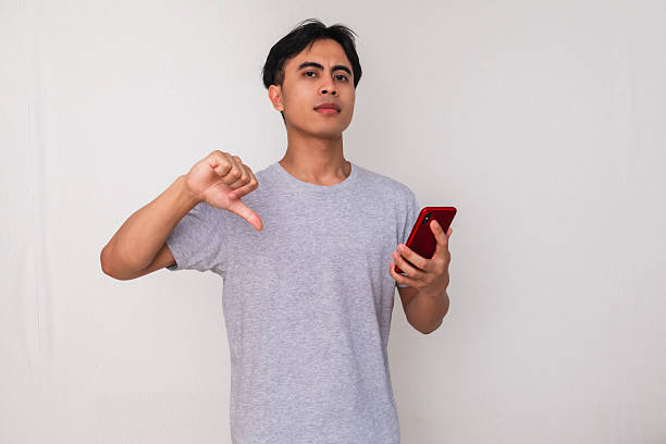istock Disappointed young Asian man showing thumbs down while holding a smartphone, isolated on white background, perfect for advertising, technology, and reaction concepts 2222688752