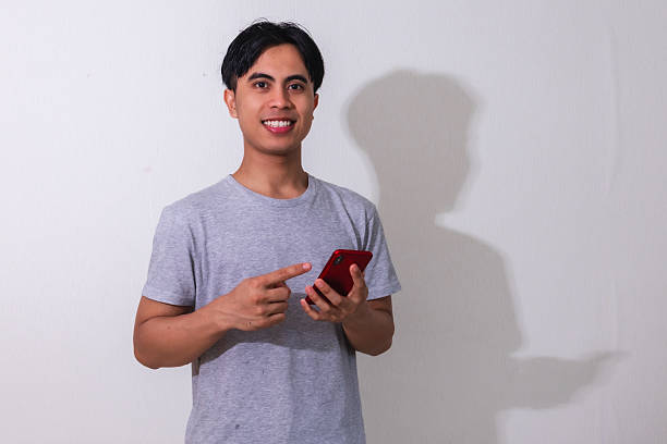 Portrait of a happy young Southeast Asian man smiling while using his smartphone on a white background, perfect for digital ads, promotions, or modern communication themes