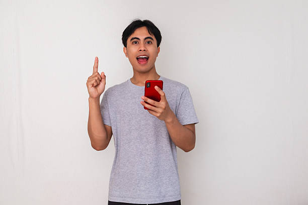 istock Excited young Asian man pointing up while holding smartphone with idea expression on isolated white background, perfect for creative app promotion or online advertisement 2222689506