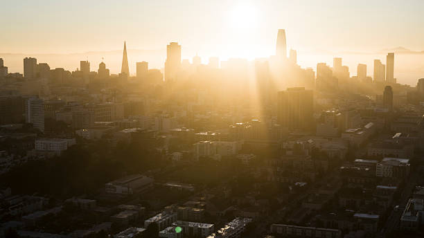 Aerial view of San Francisco skyline during sunrise with distinctive golden light creating silhouettes of skyscrapers. Prominent landmarks include Transamerica Pyramid and Salesforce Tower visible through morning haze. Sunlight streams across downtown buildings and residential areas, creating dramatic light and shadow contrast. Mission District buildings visible in foreground with downtown financial district in background. Distant hills and bay area visible through morning fog creating layered depth effect.