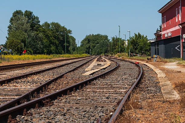 Converging railway tracks stretch into the distance at a quiet, rural train station under clear blue skies. A red building lines the platform on the right, while green vegetation and trees frame the scene on the left. The image evokes a sense of travel, waiting, and timeless infrastructure.