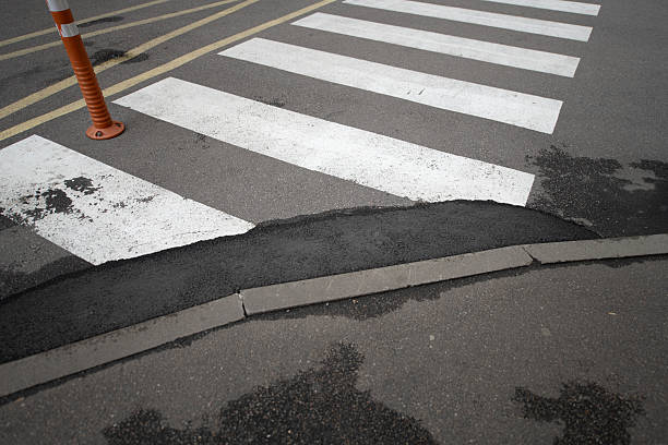 Workers are performing maintenance on a pedestrian crosswalk where the surface has cracked, revealing potholes. Traffic cones are placed nearby to alert drivers during repairs.