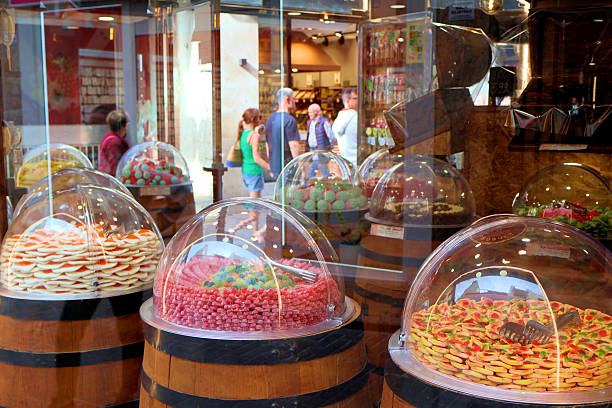 Benidorm, Spain - 2024.04.27. - A colorful candy display is showcased in a shop window, featuring a variety of sweets under transparent domes sitting atop wooden barrels. Brightly colored gummies, cookies, and other confections are invitingly arranged, drawing the eye. In the background, shoppers stroll by, adding a lively, communal atmosphere to this delightful scene.