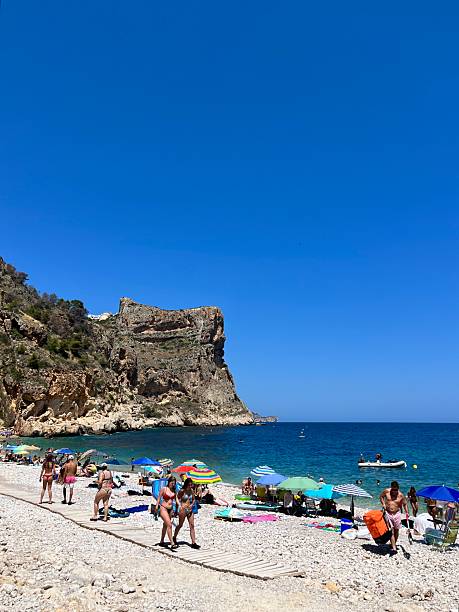 Cala Moraig, Benitachell, Javea, Valencia, Spain - June 28, 2025: Tourists walking on a wooden walkway on Cala Moraig beach in Benitachell, Spain, during a hot summer day, with the Morro Falquí cliff in the background
