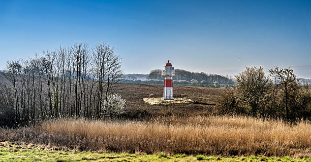 A red and white lighthouse stands on an open rural countryside, surrounded by trees, grasses, and calm vistas under a bright blue sky.