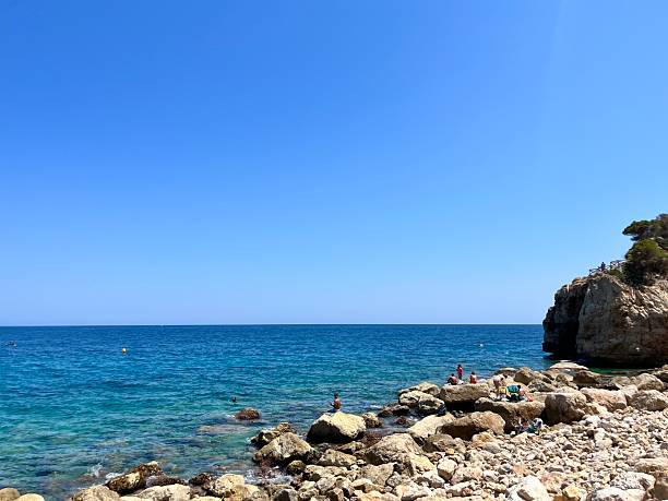 Tourists are swimming and sunbathing at Cala Moraig beach, a beautiful cove with crystal-clear turquoise waters located in Benitachell, Alicante, Spain, during a sunny summer day