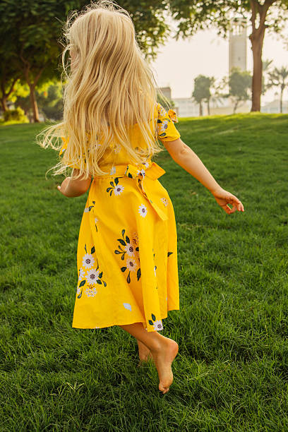A young girl with long blonde hair wearing a yellow dress, enjoying time outdoors on a sunny day. Her bare feet touch the lush green grass in a picturesque park, surrounded by trees and scenic nature, capturing a carefree and joyful moment. The photo emphasizes the beauty of childhood and the serenity of outdoor leisure.