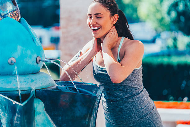 Woman in athletic wear cools off at a public fountain, splashing water on her neck after a workout on a sunny day. The vibrant outdoor setting and sports armband highlight an active, healthy lifestyle and the importance of hydration and recovery.