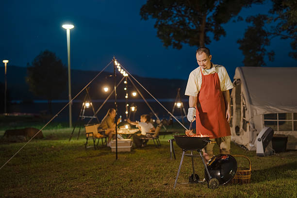 A man in an apron is standing next to a grill. There are several people around him, including a woman and two children