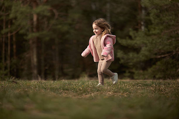 Sweet, smiling little girl in a pink jacket runs across the grass in the park.