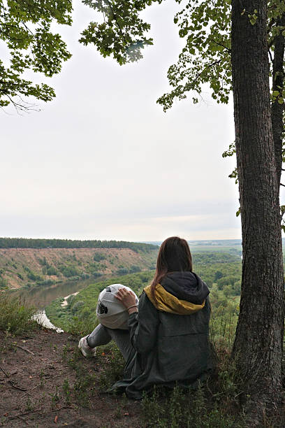 Young woman with motorcycle gear resting under a tree on a hillside overlooking the valley. Peaceful travel break moment. For adventure blogs, landscape prints, or outdoor lifestyle themes. Sstktravel