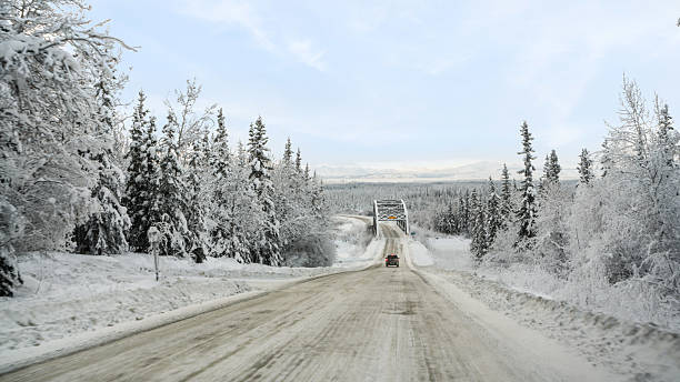 A snow-covered vehicle travels down an icy road toward a steel truss bridge that spans a frozen river in Alaska’s vast interior. Towering spruce trees coated in fresh snow line the path, creating a serene tunnel through the boreal forest. In the distance, hazy mountain peaks rise beneath a soft blue sky, adding depth and grandeur to the remote landscape. This scene captures the quiet beauty and solitude of winter travel in Alaska’s wild backcountry, where nature’s silence is broken only by the crunch of tires on packed snow.