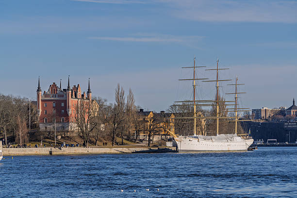 Iconic AF Chapman ship docked by the shore with castle-style building and historic houses in Skeppsholmen, Stockholm.