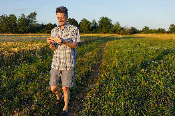 A casually dressed man with a beaming smile is actively engaged in playing a mobile game on his smartphone as he walks along a path through a vibrant green meadow and golden field. The image captures a blend of modern technology use with the tranquility of nature, highlighting leisure and digital entertainment in an outdoor setting.