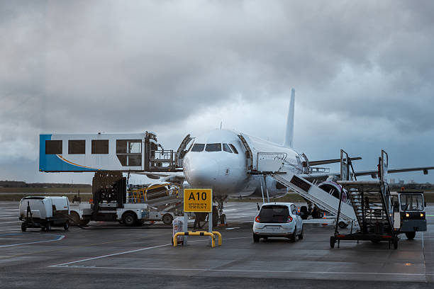 A commercial airplane is stationed at gate A10 during boarding and ground handling operations. A jet bridge is attached to the aircraft, while service vehicles and equipment surround it on a wet tarmac under a moody, overcast sky, capturing the busy and technical side of air travel.