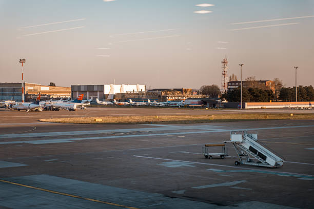 A wide view of a busy airport tarmac captured during golden hour, showing multiple commercial airplanes parked at the gate. Visible are refueling trucks, service vehicles, and various airport buildings and hangars in the background. The warm evening light creates a soft and cinematic atmosphere, ideal for illustrating global air travel, aviation logistics, and the infrastructure behind modern transportation networks.