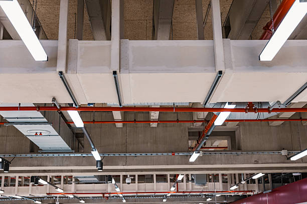 Detailed view of an airport ceiling showing exposed HVAC air ducts, fluorescent lighting, and structural beams. The image captures modern infrastructure and industrial architecture design, ideal for illustrating engineering and facility management themes.