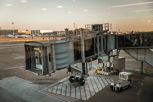 A detailed view of a covered staircase leading to a jet bridge at an airport terminal. The ground features clearly marked pedestrian walkways in blue, bordered by traffic cones and safety chains. The image highlights the functional design of modern airport infrastructure, focusing on access paths and boarding systems. Perfect for themes related to air travel, transport logistics, passenger movement, and architectural elements of airports.