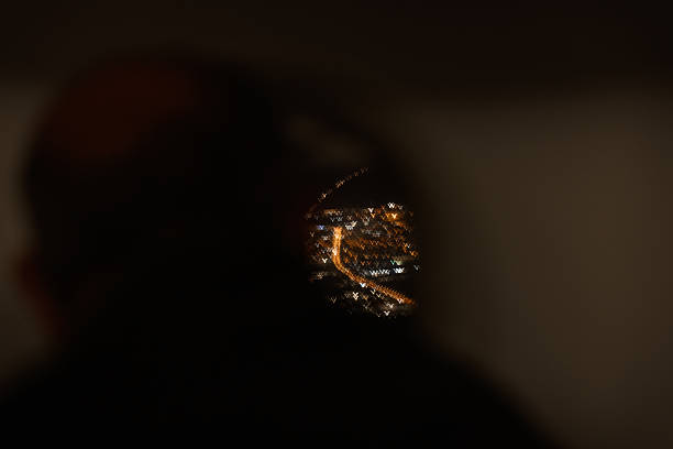 Blurry night view of a city illuminated with golden street lights, seen through an airplane window. The silhouette of a passenger is subtly visible in the foreground, evoking the experience of travel, night descent, and reflective moments during flight.