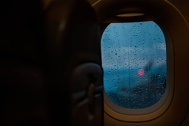 A melancholic and cinematic view through an airplane window covered with raindrops. The image captures the atmosphere of a rainy day at the airport, viewed from inside the cabin during boarding or taxiing, with soft interior lighting adding warmth.