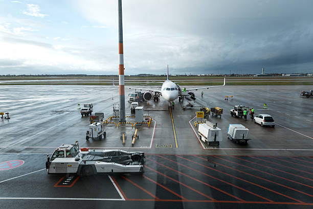 Wide-angle shot of a commercial airplane on a wet airport apron during ground operations. Ground crew members are actively working around the aircraft, preparing it for departure, while various support vehicles are parked nearby under a dramatic, overcast sky.