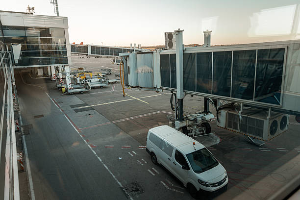 View from an airport terminal showing an empty jet bridge extending toward the tarmac, accompanied by a white airport service van and various ground handling equipment. The area is quiet and organized, reflecting early morning or late evening light. This image emphasizes transportation logistics, air travel infrastructure, and behind-the-scenes airport operations, ideal for themes of aviation, travel, and airport services.