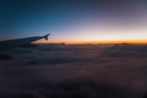 Stunning view from an airplane window showcasing the aircraft wing flying above thick clouds during a colorful sunset. The soft gradients of the sky create a dreamy and peaceful travel scene captured mid-flight.