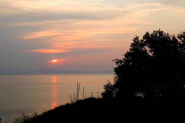 Vibrant Sunset Over A Calm Sea, With The Sun Dipping Below The Horizon And Casting A Warm Orange Glow Across The Sky And Water. Silhouetted Trees And Grassy Hill In The Foreground Add Depth And Mood To This Peaceful Nature Scene.