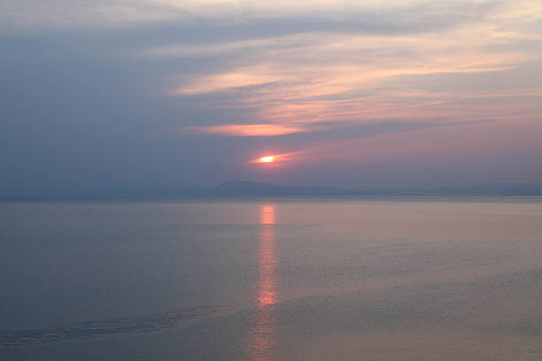 Minimalistic Sunset Over A Still Sea, With Soft Pink, Purple, And Blue Tones In The Sky. The Sun Is Setting Behind Distant Hills, Creating A Gentle Reflection On The Water Surface. The Image Evokes A Peaceful, Dreamlike Mood.