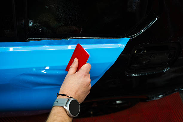 istock Detailing expert uses red squeegee wrapper to flatten blue PPF wrap on car surface. Vehicle wrap detailing involves squeegee strokes for seamless, wrinkle-free adhesion. 2222786858