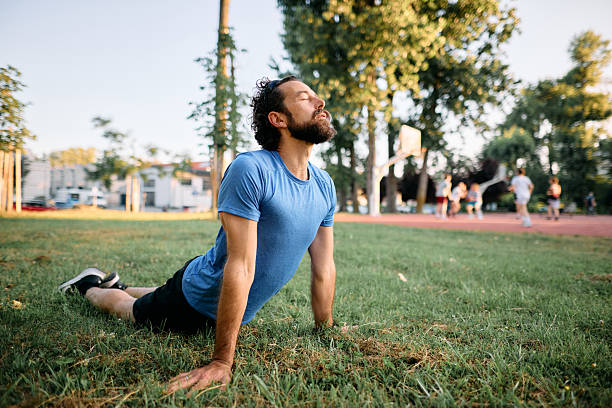 Mid adult man is stretching his body in a park on a sunny day, performing the upward facing dog asana, a yoga posture, while in the background some people are playing basketball