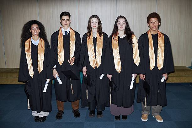 Group of five multiethnic teenagers standing in graduation gowns holding diplomas, facing camera with cheerful expressions, representing university students at commencement ceremony