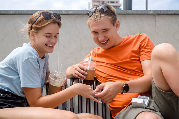 A guy puts an engagement ring on his girlfriend's finger while they are sitting on a city bench with cocktails in their hands. Close-up
