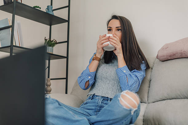istock Young woman drinking coffee while watching tv on sofa at home 2222835461