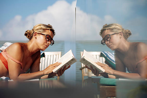 A blonde-haired woman in a coral swimsuit reads a book near a swimming pool