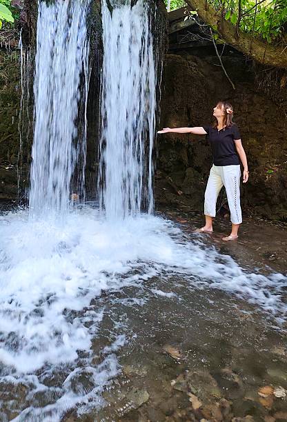 Female swimmer in blue bikini walking through waterfall spray with arms outstretched. Natural forest waterfall setting with sunlight filtering through trees creating dramatic lighting. Joyful outdoor summer recreation moment embracing nature in Norwegian wilderness. Refreshing water experience and adventure lifestyle.