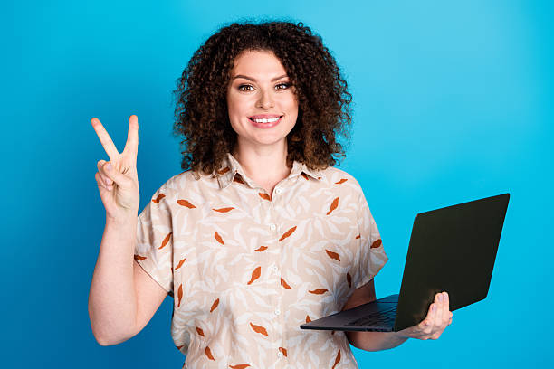 istock Smiling young woman holding a laptop while making a peace gesture against a bright blue background 2222847279