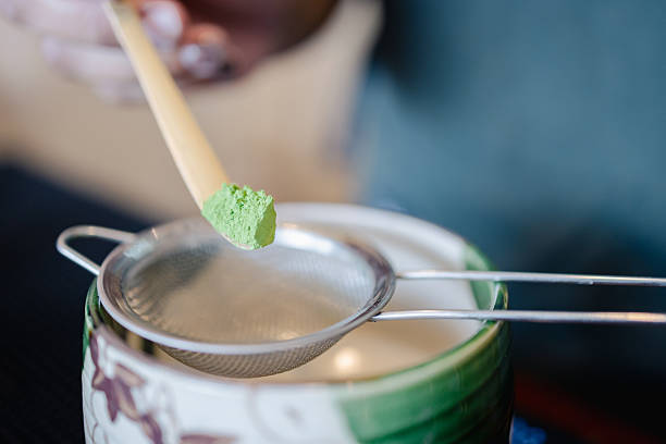 Green tea powder held by wooden spoon above metal strainer and ceramic cup with floral pattern in soft focus background creating calm mood for tea preparation