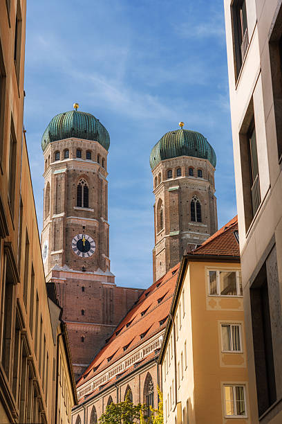 View of the iconic Frauenkirche cathedral towers in Munich, Germany, prominently framed between city buildings under a clear blue sky.