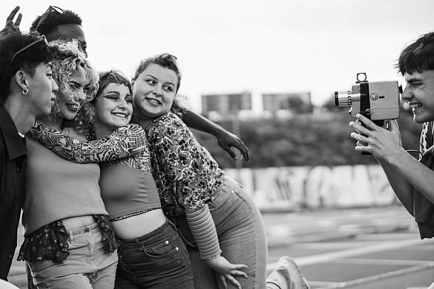 Young diverse people having fun with old vintage video camera outdoor - Main focus on blond girl face - Black and white editing