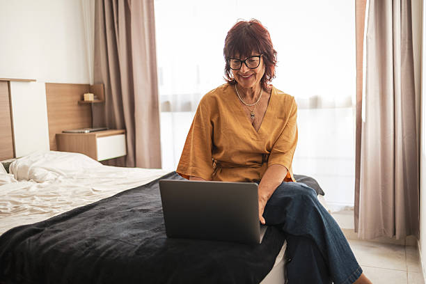 An elderly woman wearing glasses and a yellow blouse is seated on a bed using a laptop computer. The hotel room is bright, featuring a comfortable interior and a relaxed, peaceful ambiance.