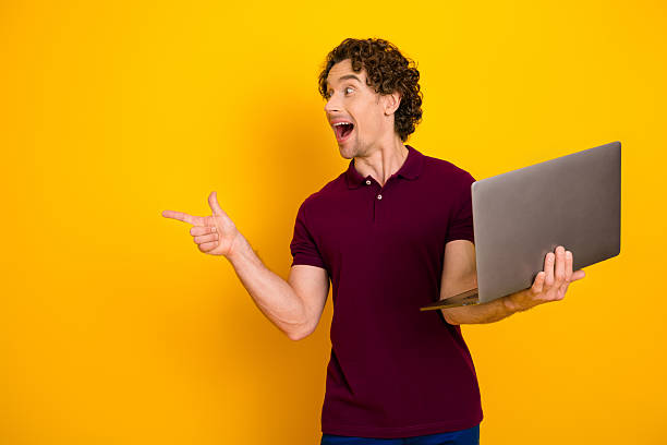 istock Cheerful man with curly hair showcasing a laptop while pointing energetically against a vibrant yellow background 2222862854