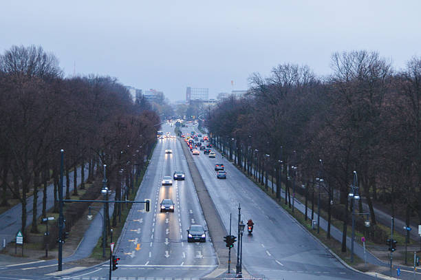 Rainy evening view from the Victory Column in Berlin, Germany. Cars and a motorbike move along a wet boulevard lined with bare trees, with city buildings in the distance.