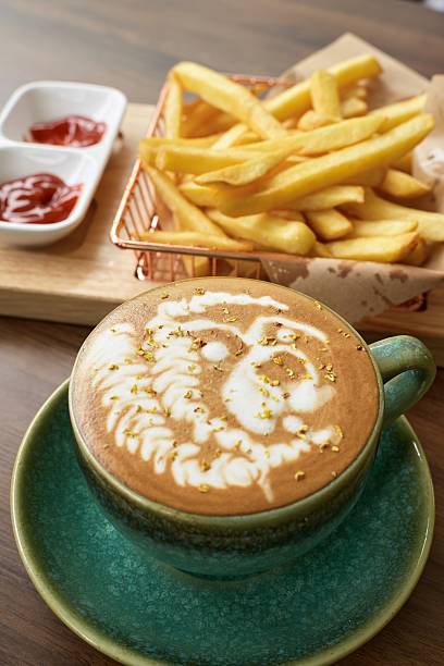 A cup of coffee with a gold leaf design on it sits on a table next to a basket of french fries