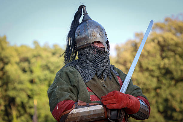 Medieval mustachioed knight in armor with sword, standing near wooden castle at dusk, historical reenactment.