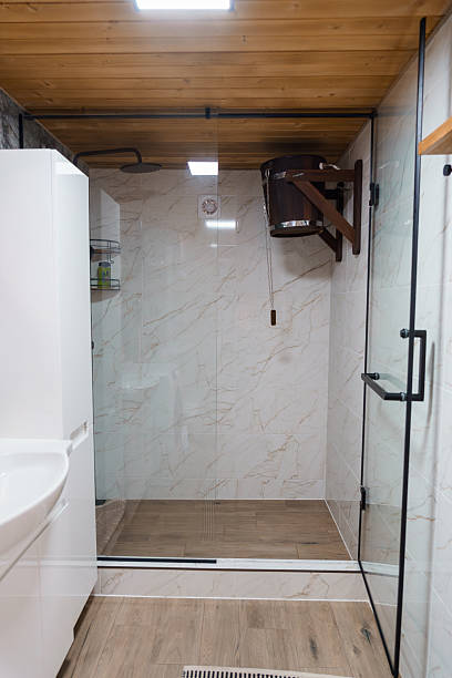 A bright, clean shower room showcases a glass-enclosed walk-in shower with marble tile and a wooden floor, complemented by a cedar ceiling and dark wood sauna heater