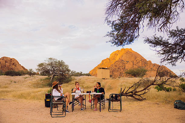 Front view of three female friends resting by the picnic table with food and drinks surrounded by majestic cliffs and desert area of Spitzkoppe during bright sunset