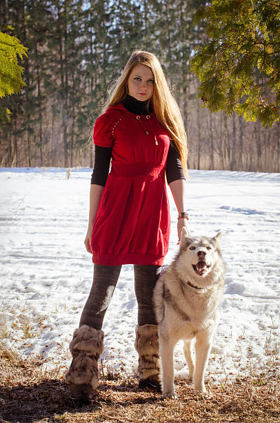 Young woman with long blonde hair stands beside dog in snowy forest. Red dress adds contrast to white surroundings. Sunlight filters through trees, creating warm ambiance