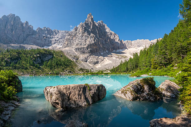 A breathtaking turquoise alpine lake lies beneath a towering rocky mountain peak, surrounded by dense green pine forests. Large boulders emerge from the crystal-clear water, reflecting the rugged landscape and blue sky above.